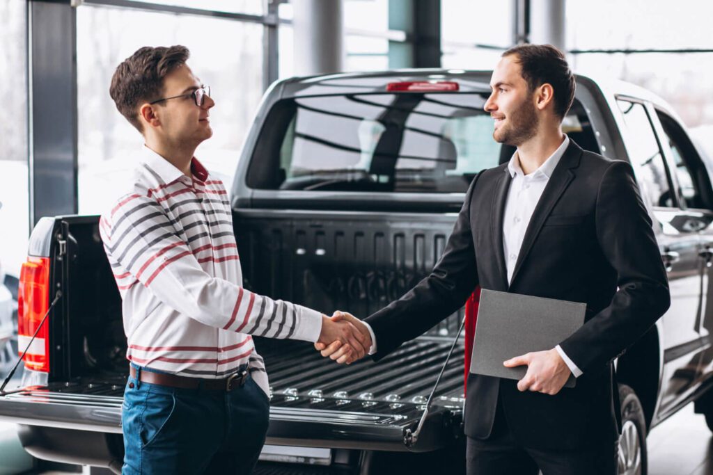 Two men shaking hands near a pickup truck, one holding a folder.